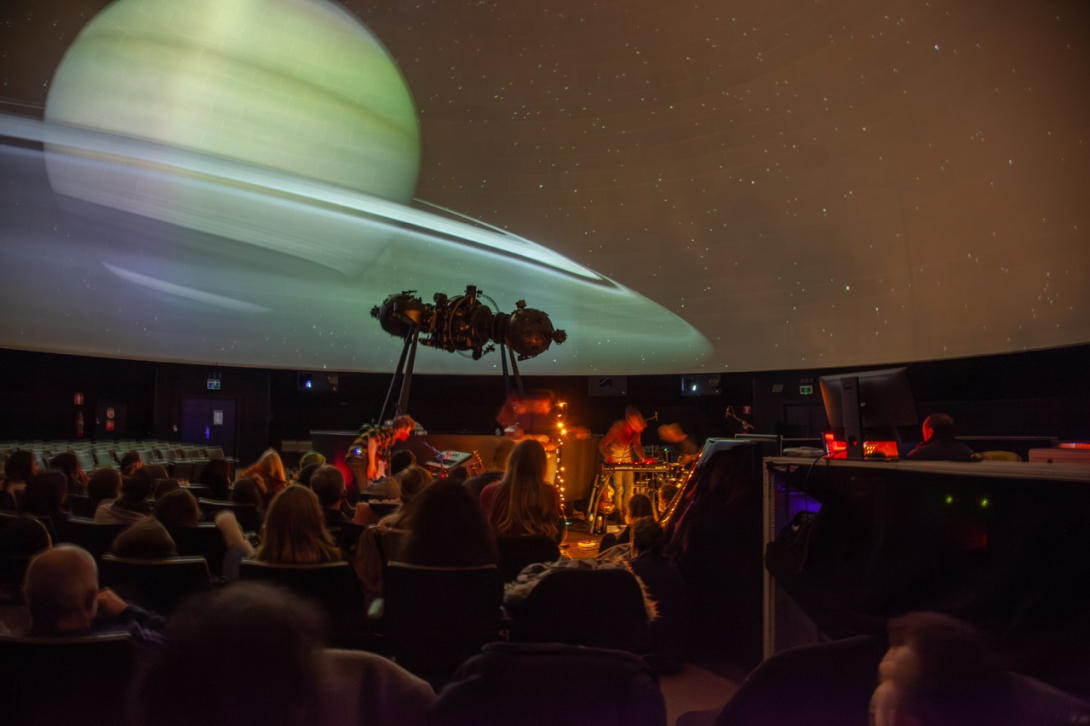 View of the dome in the Planetarium with musicians in front of an image of a planet and the public watching.